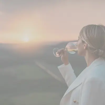 Frau steht vor einem Aussichtspunkt und schaut in den Sonnenuntergang. Sie trinkt einen Schluck Weisswein.
