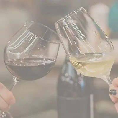 Zwei Personen stossen mit einem Rotwein- und einem Weissweinglas an, im Hintergrund steht eine Weinflasche.