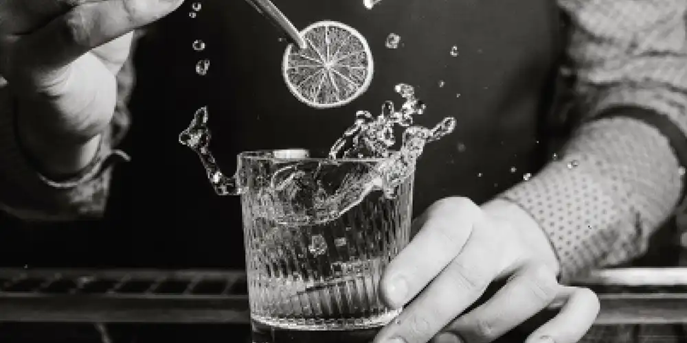 Schwarz-Weiss-Bild von einer Hand, die eine getrocknete Orangenscheibe in ein tiefes Glas gibt.